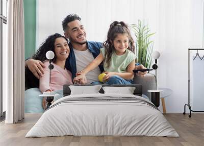 Family Weekend. Cheerful arabic parents and little daughter watching tv at home Wall mural