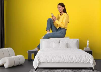 Female Using Smartphone Texting Sitting On Cube Over Yellow Background Wall mural