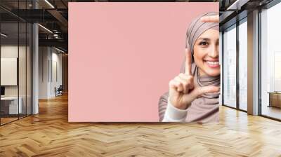 Frame your beauty. Smiling muslim girl in hijab making frame with fingers, capturing moment, posing over pink background with empty space Wall mural