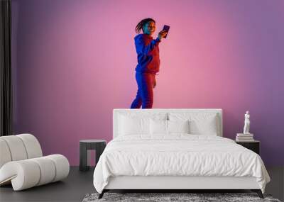 Full body length of young sporty black woman in sportswear jumping and using mobile cellphone on pink neon background Wall mural