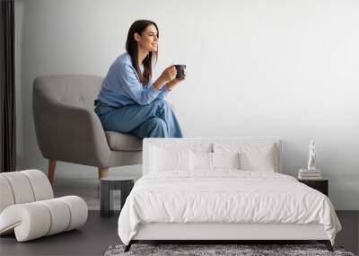 Full length of young woman drinking hot coffee in cozy armchair against white studio wall, banner with free space Wall mural