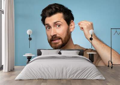 Funny middle aged bearded man cleaning ears with cotton swab, doing daily hygiene routine, blue studio background Wall mural