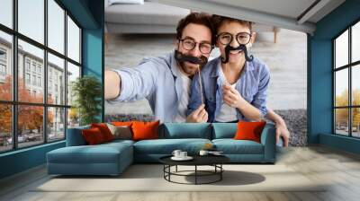 Funny selfie. Happy father and son taking selfie, holding fake moustache on sticks and smiling at camera Wall mural