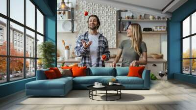 Glad caucasian millennial male with stubble juggles tomatoes, blonde lady prepares salad Wall mural