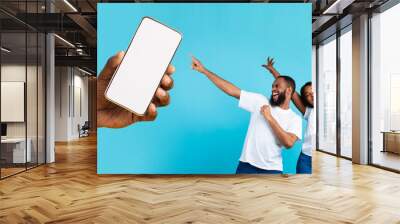 Great Offer. Cheerful African American couple dancing and pointing at giant cell phone showing blank space for mock up, celebrating win, hand holding white empty screen, blue studio background wall Wall mural