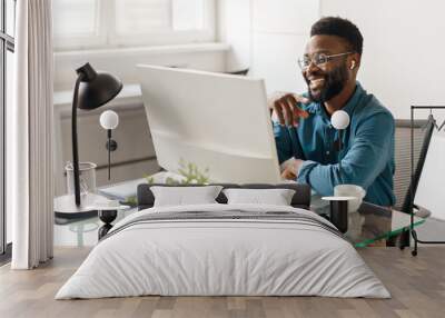 Handsome black male entrepreneur sitting at desk in office, looking at computer monitor and smiling, having video call Wall mural