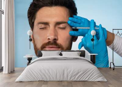 Handsome middle aged bearded man getting under eye injections in beauty clinic, blue studio background, closeup Wall mural