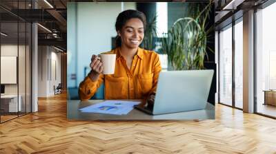 Happy african american businesswoman working on laptop and drinking coffee, sitting at workplace in office Wall mural