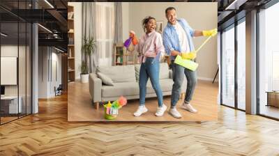 Happy african american spouses having fun with mop and detergent bottle, singing while cleaning house Wall mural