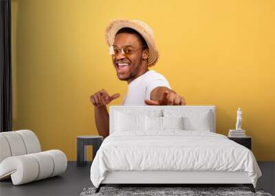 Happy black guy in casual summer outfit, straw hat and sunglasses pointing at you on yellow studio background Wall mural