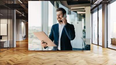 Happy businessman talking on smartphone and checking papers, standing in modern office interior, free space Wall mural