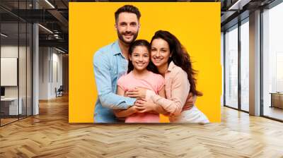 Happy european parents hugging their pretty daughter and smiling to camera on yellow background, studio shot Wall mural