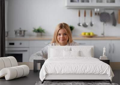 Happy european pretty adolescent blonde girl in casual sits at table with glass of water in minimalist kitchen Wall mural