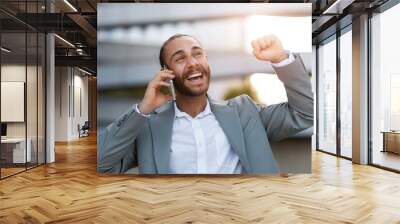 Happy excited young businessman talking on cellphone outdoors and celebrating success Wall mural