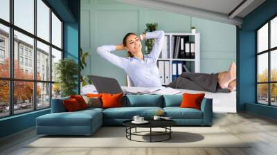 Happy female entrepreneur having time-out at workplace, dreaming while sitting on armchair and putting feet on table Wall mural