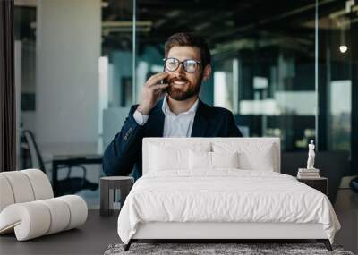 Happy handsome businessman working on laptop and having phone conversation, sitting in office and smiling Wall mural