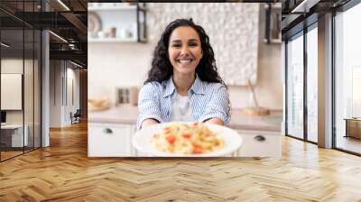 Happy latin woman showing spaghetti in the dish, stretching meal to camera and smiling Wall mural