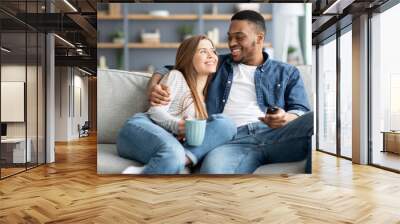 Happy loving interracial couple relaxing at home, watching tv and drinking coffee Wall mural