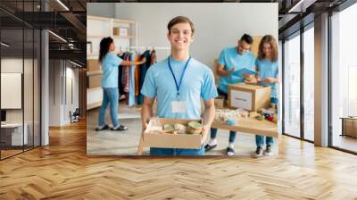 Happy male activist volunteer in blue uniform holding cardboard box with food for donation, working in charity center Wall mural