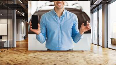 Happy man showing new car key and smartphone with blank screen standing in automobile dealership center, mock up Wall mural