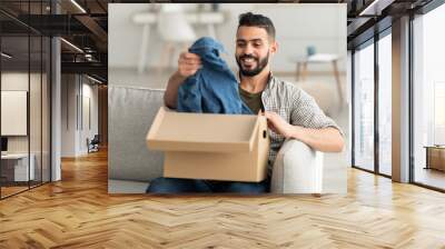 Happy millennial Arab guy unboxing carton parcel, satisfied with successful shopping, taking out new clothes at home Wall mural