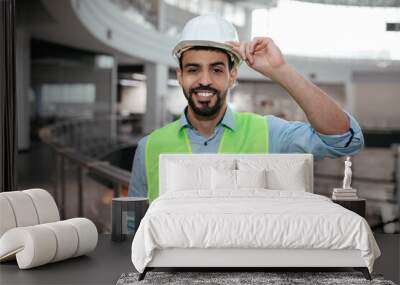 Happy millennial middle eastern builder engineer with beard in protective clothing touches hard hat with hand Wall mural