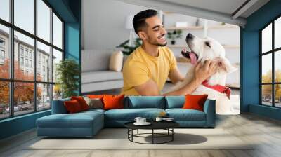 Happy young Arab man stroking his adorable dog in living room. Human animal friendship concept Wall mural