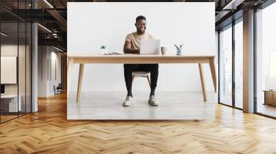 Happy young black man using laptop computer for online work at table in home office, free space Wall mural