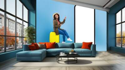 Happy young black woman sitting on suitcase and pointing at smartphone with mockup for online travel agency website Wall mural