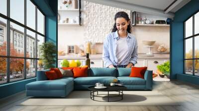 Happy young lady making notes on table with organic vegetables, cooking dinner in light kitchen interior Wall mural