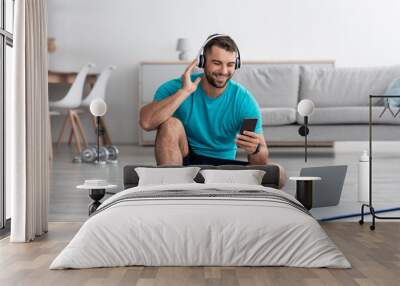 Happy young muscular european guy in headphones sits on mat with bottle of water and pc, typing on phone Wall mural