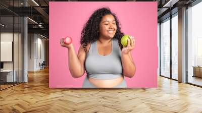 Healthy diet concept. Happy black body positive woman holding dumbbell and apple, enjoying fresh fruit and workout Wall mural