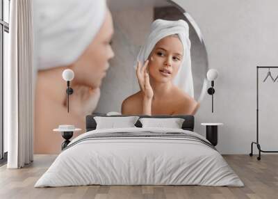 Home beauty routine. Young woman applying moisturising cream on face looking at miror in bathroom, free space Wall mural