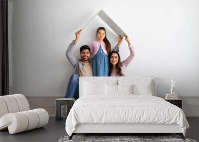 Housing for family. Young eastern parents joining hands making symbolic roof above child, sitting on floor near white wall Wall mural