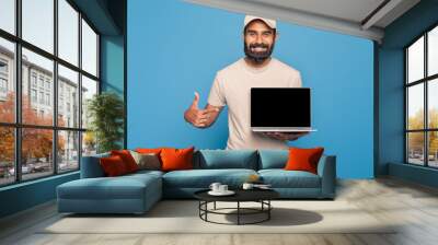 Indian man is standing while holding a laptop with black screen in one hand and giving a thumbs up gesture with the other hand Wall mural