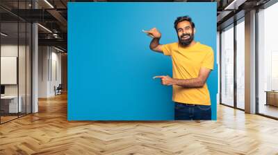 Indian man wearing a yellow shirt is pointing at an object or direction outside the frame of the image. His arm is extended, and his index finger is directed towards the unseen subject, copy space Wall mural