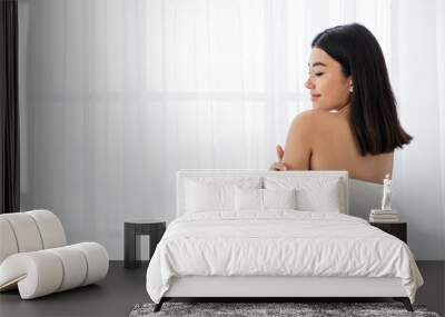 Joyful asian lady in towel touching her skin while standing by window at home. Young woman wrapped in white bath towel enjoying her soft skin after morning shower, panorama with copy space Wall mural