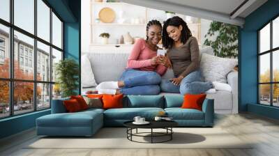 Joyful black girlfriend sitting on sofa and using smartphone Wall mural