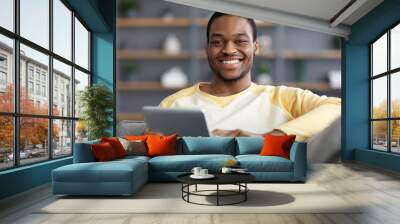 Joyful black guy resting with digital tablet on couch Wall mural