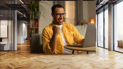 Joyful millennial black man with laptop celebrating online win or work success at home Wall mural