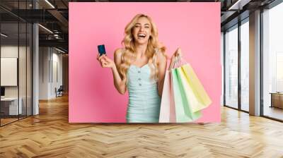 Laughing young woman with long blonde hair smiles brightly at the camera. She holds a credit card in her left hand and several colorful shopping bags in her right Wall mural