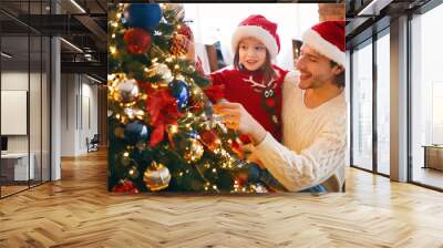 Little girl in red Santa hat enjoying Christmas tree with her father Wall mural