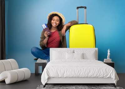 Lovely young black woman holding passport and plane tickets, sitting near bright yellow suitcase on blue background Wall mural