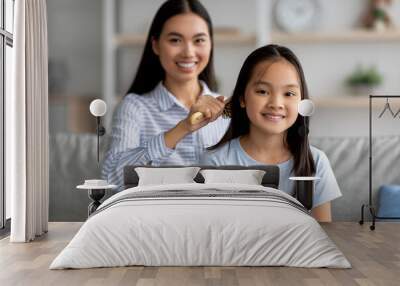 Loving asian mother combing her daughter hair with wooden brush while sitting together on sofa at home Wall mural