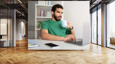 Man in a green t-shirt working on a laptop at a white desk, drinking coffee from a blue mug Wall mural