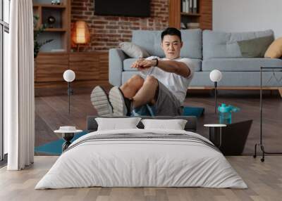 Mature korean man doing exercises for abs on mat on floor in living room interior, watching workout on laptop computer Wall mural