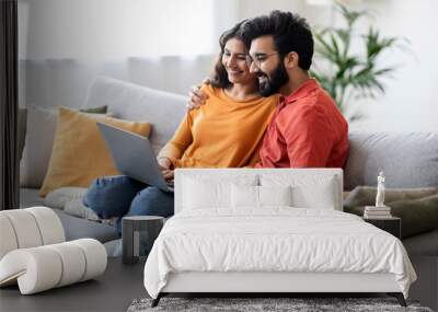 Millennial Happy Indian Couple Relaxing With Laptop On Couch At Home Wall mural