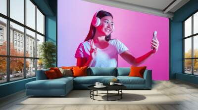 Music and selfie. Smiling girl in headphones shows peace sign and makes photo Wall mural