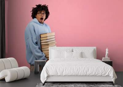 Nerdy black teenage student feeling shocked over pile of homework, holding big stack of books on pink background Wall mural