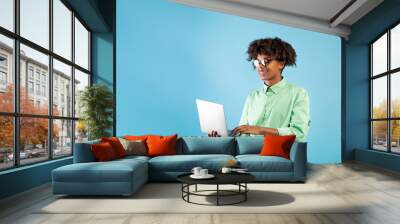 Online education programs. Black teen guy using laptop while standing over blue background, studio shot, free space Wall mural